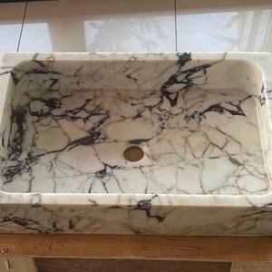 May include: A white marble sink with black veining. The sink is rectangular and has a drain hole in the center. It is sitting on a wooden base.