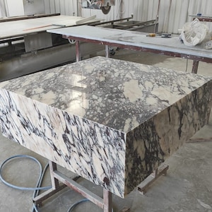 May include: A large, rectangular block of marble with a polished surface. The marble features a complex pattern of white, beige, and dark gray veining. The block is supported by a metal stand, suggesting it is a countertop or slab for construction.