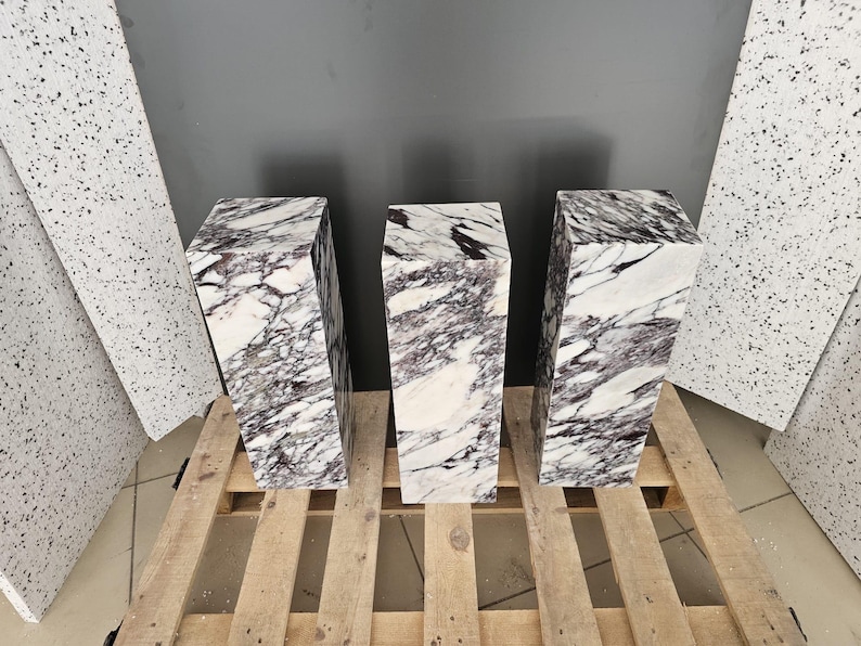 May include: Three white marble pedestals with purple veining. The pedestals are arranged in a row on a wooden pallet.