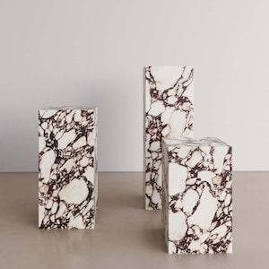 May include: Three white marble pedestals with brown veining. The pedestals are different sizes and are arranged in a row.