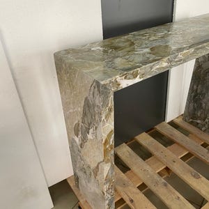 Pu&ograve; includere: Un tavolo consolle lungo e stretto in marmo verde e marrone con finitura lucida. Il tavolo &egrave; sostenuto da un'unica e spessa gamba in marmo a ciascuna estremit&agrave;.