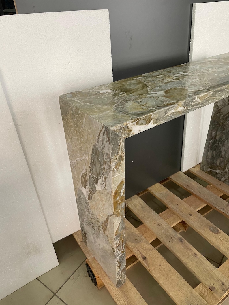 Pu&ograve; includere: Un tavolo consolle lungo e stretto in marmo verde e marrone con finitura lucida. Il tavolo &egrave; sostenuto da un'unica e spessa gamba in marmo a ciascuna estremit&agrave;.