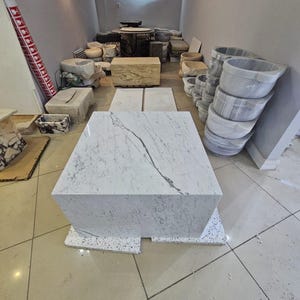 May include: A large, white marble cube-shaped coffee table with gray veining. The table is displayed in a showroom with other stone products, including sinks and tables. The floor is tiled.