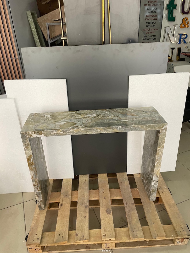 Pu&ograve; includere: Un tavolino da console in marmo verde chiaro con un piano rettangolare e un design semplice e moderno. Il tavolo &egrave; appoggiato su un pallet di legno.