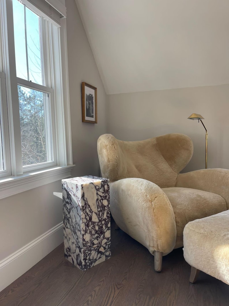 May include: A white and purple marble side table with a fluffy white sheepskin armchair and ottoman in a room with a large window and a framed picture on the wall.