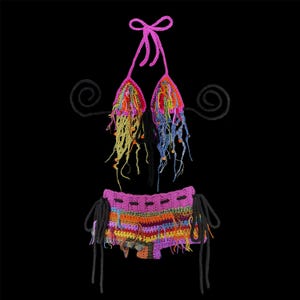 May include: A two-piece crochet set consisting of a pink halter top and matching shorts. The top features dangling multicolored yarn fringe and thin pink ties. The shorts have horizontal stripes in orange, yellow, purple, and blue with black side ties.