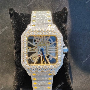 May include: A luxury wristwatch with a square face and a silver strap, heavily encrusted with sparkling gemstones. The watch has a gold-coloured frame and a blue second hand. The watch is displayed on a black velvet surface.