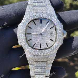 May include: A luxury wristwatch with a silver strap and face, entirely covered in sparkling gemstones. The watch has a round face with black markers and hands, and a date display. The watch is held by a gloved hand.