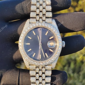 May include: A luxury wristwatch with a dark blue face, surrounded by a bezel and band encrusted with sparkling stones. The watch features a date display. The watch is set against a blurred background. The brand name "Diamond Blings" is visible.