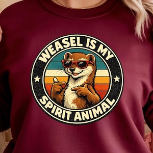 Puede incluir: Sudadera burdeos con un gráfico circular. El gráfico presenta una comadreja de dibujos animados con gafas de sol y el texto "WEASEL IS MY SPIRIT ANIMAL". El diseño incluye una puesta de sol retro y acentos de estrellas. Un círculo rojo dice "DESCARGA INSTANTÁNEA".