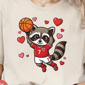 May include: Beige t-shirt featuring a cartoon raccoon in a red basketball jersey with the number 7, holding a basketball. The design includes red and pink hearts. The words "INSTANT DOWNLOAD" are in a red circle.