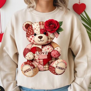 May include: A beige sweatshirt featuring a patchwork teddy bear design. The bear has a red heart with the word "LOVE" and the words "HUGS" and "KISSES" on its paws. A red rose adorns the bear's head, perfect for Valentine's Day.
