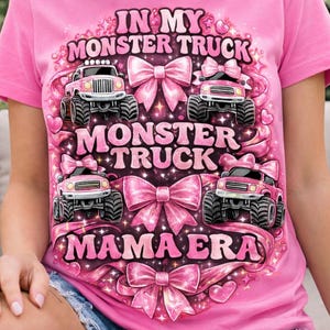 May include: Pink t-shirt with a graphic design featuring pink monster trucks, bows, and the text "IN MY MONSTER TRUCK," "MONSTER TRUCK," and "MAMA ERA." The design is surrounded by hearts and floral accents.