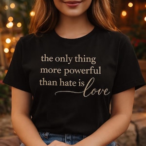 Puede incluir: Camiseta negra con el texto "the only thing more powerful than hate is love" en escritura beige claro. La camiseta se usa con pantalones cortos de mezclilla. El fondo está borroso con luces cálidas.