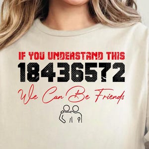 Könnte beinhalten: Cremefarbenes T-Shirt mit dem Text "IF YOU UNDERSTAND THIS 18436572 We Can Be Friends" in Rot und Schwarz. Das Shirt hat auch eine kleine Grafik von zwei sich umarmenden Figuren. Auf der Unterseite des Shirts steht "USMODERNLUXURY".