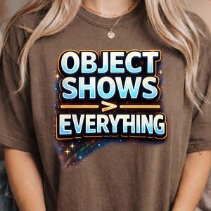 May include: A brown t-shirt featuring the text "OBJECT SHOWS > EVERYTHING" in a blue and yellow design. The shirt has a relaxed fit, suitable for casual wear. The design includes a starry background.
