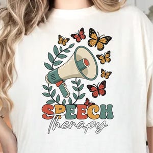 May include: Cream-colored t-shirt featuring a retro design with a megaphone, butterflies, and the words "SPEECH Therapy." The design includes green leaves and colorful text. The shirt is a casual, comfortable style.
