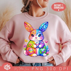 Może przedstawiać: Jasnoróżowa bluza z kolorową, szczegółową ilustracją zajączka wielkanocnego. Zajączek jest ozdobiony żywymi wzorami, trzyma jajko i jest otoczony kwiatami i sercami. Słowa "INSTANT DOWNLOAD" są w czerwonym okręgu.