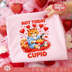 May include: A pale pink t-shirt featuring a cartoon ginger cat in a blue robe, holding a remote and a bowl of snacks. The text "NOT TODAY CUPID" is printed in red. Red heart balloons and a gift box are also shown.
