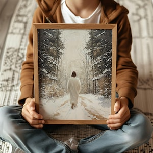 May include: A framed painting of a person in a white robe walking down a snowy path in a forest. The path is lined with trees on either side and the sky is a light blue. The painting is in a wooden frame.