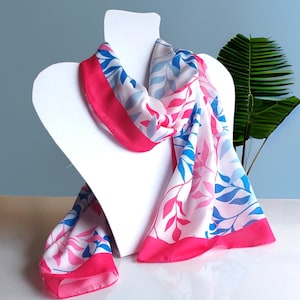 May include: A pink and blue floral patterned scarf with a white background. The scarf is folded and draped over a white mannequin head.