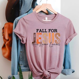 Puede incluir: Camiseta de color malva con la frase "FALL FOR JESUS He Never Leaves" en letras negras, naranjas y beige. La camiseta est&aacute; colgada en una percha de madera, con una chaqueta vaquera y un bolso de cuero marr&oacute;n al fondo.