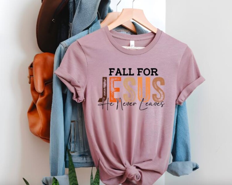 Puede incluir: Camiseta de color malva con la frase "FALL FOR JESUS He Never Leaves" en letras negras, naranjas y beige. La camiseta est&aacute; colgada en una percha de madera, con una chaqueta vaquera y un bolso de cuero marr&oacute;n al fondo.