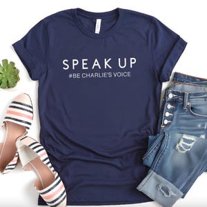 Puede incluir: Camiseta azul marino con las palabras "SPEAK UP" y el hashtag "#BE CHARLIE'S VOICE" en blanco. La imagen tambi&eacute;n presenta vaqueros azules desgastados, bailarinas a rayas y una peque&ntilde;a planta suculenta.