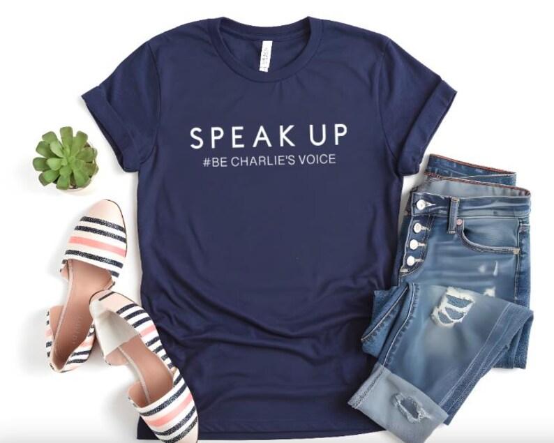 Puede incluir: Camiseta azul marino con las palabras "SPEAK UP" y el hashtag "#BE CHARLIE'S VOICE" en blanco. La imagen tambi&eacute;n presenta vaqueros azules desgastados, bailarinas a rayas y una peque&ntilde;a planta suculenta.