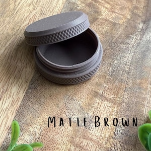 May include: A matte brown container with a lid slightly ajar, revealing the interior. The lid and base have a textured, ridged design. The text "MATTE BROWN" is written in black below the container.