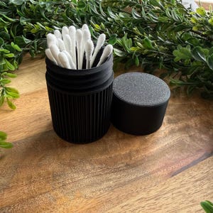 May include: A black, cylindrical container with a textured lid, filled with white cotton swabs. The container has vertical grooves and sits on a wooden surface, with green foliage in the background. The lid is next to the container.