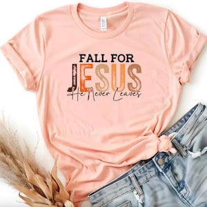 Puede incluir: Una camiseta color melocot&oacute;n con las palabras "FALL FOR JESUS He Never Leaves" en una mezcla de letras marrones, naranjas y negras. La camiseta se combina con pantalones cortos de mezclilla. Un manojo de hierba ornamental seca est&aacute; en la esquina inferior izquierda.