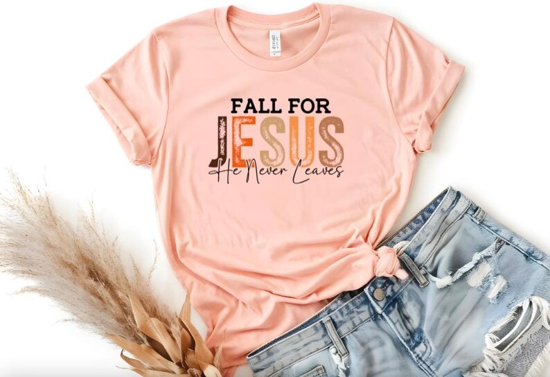 Puede incluir: Una camiseta color melocot&oacute;n con las palabras "FALL FOR JESUS He Never Leaves" en una mezcla de letras marrones, naranjas y negras. La camiseta se combina con pantalones cortos de mezclilla. Un manojo de hierba ornamental seca est&aacute; en la esquina inferior izquierda.