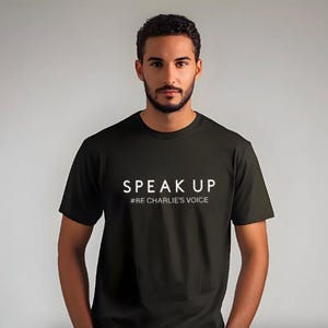 Puede incluir: Una camiseta negra con las palabras "SPEAK UP" en letras blancas. Debajo, la frase "#BE CHARLIE'S VOICE" en una fuente m&aacute;s peque&ntilde;a. La camiseta es de manga corta y parece ser de algod&oacute;n.