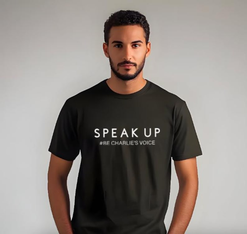 Puede incluir: Una camiseta negra con las palabras "SPEAK UP" en letras blancas. Debajo, la frase "#BE CHARLIE'S VOICE" en una fuente m&aacute;s peque&ntilde;a. La camiseta es de manga corta y parece ser de algod&oacute;n.