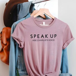 Puede incluir: Una camiseta de color malva con las palabras "SPEAK UP" impresas en negro. Debajo del texto est&aacute; "#BE CHARLIE'S VOICE". La camiseta est&aacute; colgada en una percha de madera con una chaqueta vaquera y una mochila de cuero marr&oacute;n en el fondo.