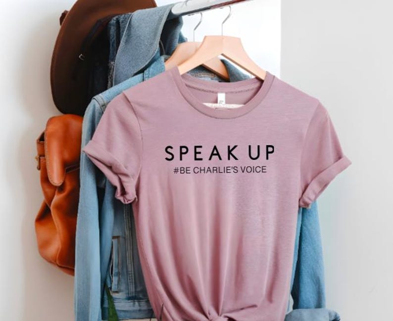 Puede incluir: Una camiseta de color malva con las palabras "SPEAK UP" impresas en negro. Debajo del texto est&aacute; "#BE CHARLIE'S VOICE". La camiseta est&aacute; colgada en una percha de madera con una chaqueta vaquera y una mochila de cuero marr&oacute;n en el fondo.