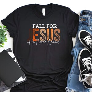 Puede incluir: Camiseta negra con el texto "FALL FOR JESUS He Never Leaves" en blanco, marr&oacute;n y naranja. La camiseta se combina con vaqueros azules y zapatillas negras con cordones blancos. Una peque&ntilde;a planta en maceta y un cuaderno tambi&eacute;n est&aacute;n en la imagen.