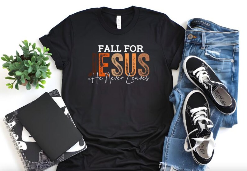 Puede incluir: Camiseta negra con el texto "FALL FOR JESUS He Never Leaves" en blanco, marr&oacute;n y naranja. La camiseta se combina con vaqueros azules y zapatillas negras con cordones blancos. Una peque&ntilde;a planta en maceta y un cuaderno tambi&eacute;n est&aacute;n en la imagen.
