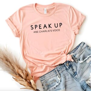 Puede incluir: Una camiseta color melocot&oacute;n con las palabras "SPEAK UP" y "#BE CHARLIE'S VOICE" impresas en negro. La camiseta se combina con unos shorts vaqueros de color azul claro. Un manojo de hierba ornamental seca se coloca a la izquierda de la camiseta.