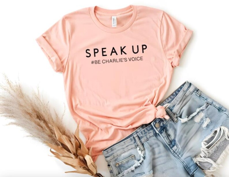 Puede incluir: Una camiseta color melocot&oacute;n con las palabras "SPEAK UP" y "#BE CHARLIE'S VOICE" impresas en negro. La camiseta se combina con unos shorts vaqueros de color azul claro. Un manojo de hierba ornamental seca se coloca a la izquierda de la camiseta.