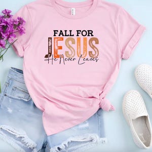 Puede incluir: Una camiseta rosa claro con el texto "FALL FOR JESUS He Never Leaves" en negro, naranja y marr&oacute;n. La camiseta se combina con pantalones cortos de mezclilla, zapatos blancos y flores moradas.