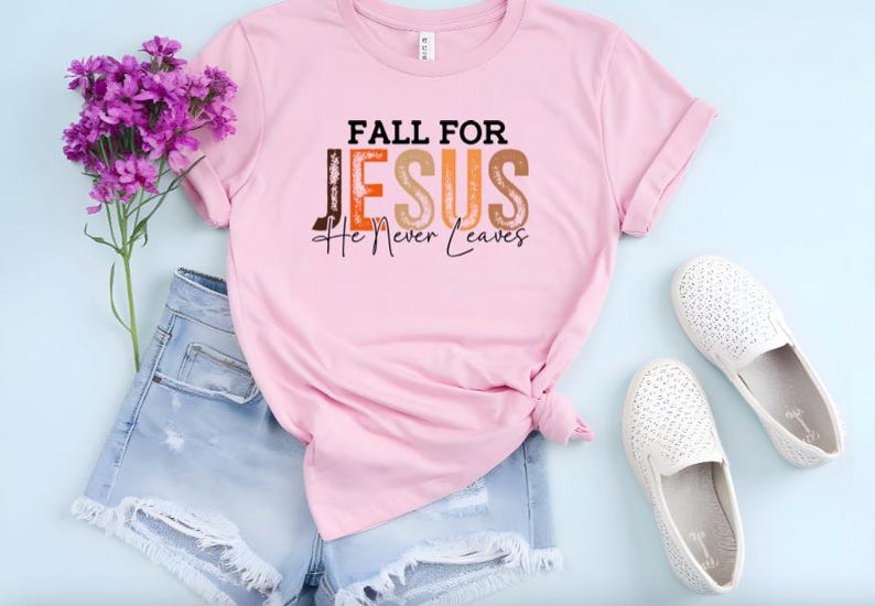 Puede incluir: Una camiseta rosa claro con el texto "FALL FOR JESUS He Never Leaves" en negro, naranja y marr&oacute;n. La camiseta se combina con pantalones cortos de mezclilla, zapatos blancos y flores moradas.