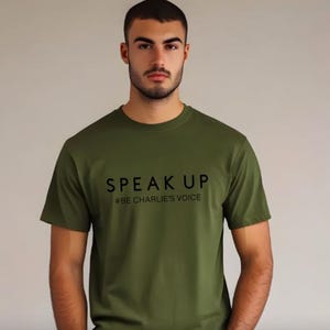 Puede incluir: Camiseta verde oliva con las palabras "SPEAK UP" y el hashtag "#BE CHARLIE'S VOICE" impresos en negro. La camiseta es de cuello redondo y tiene mangas cortas. El fondo es de un color neutro s&oacute;lido.