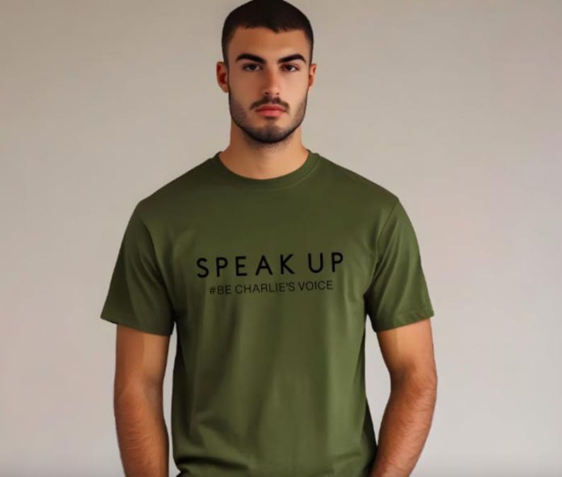 Puede incluir: Camiseta verde oliva con las palabras "SPEAK UP" y el hashtag "#BE CHARLIE'S VOICE" impresos en negro. La camiseta es de cuello redondo y tiene mangas cortas. El fondo es de un color neutro s&oacute;lido.