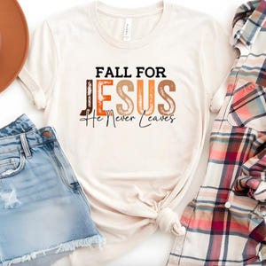 Puede incluir: Camiseta color crema con el texto "FALL FOR JESUS He Never Leaves" en marr&oacute;n, naranja y negro. La camiseta se combina con pantalones cortos de mezclilla, un sombrero marr&oacute;n y una camisa a cuadros.