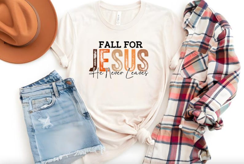 Puede incluir: Camiseta color crema con el texto "FALL FOR JESUS He Never Leaves" en marr&oacute;n, naranja y negro. La camiseta se combina con pantalones cortos de mezclilla, un sombrero marr&oacute;n y una camisa a cuadros.