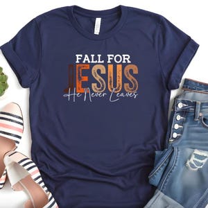 Puede incluir: Camiseta azul marino con el texto "FALL FOR JESUS He Never Leaves" en blanco, naranja y marr&oacute;n. La camiseta se combina con vaqueros azules y bailarinas a rayas. Tambi&eacute;n se ve una peque&ntilde;a planta suculenta verde.