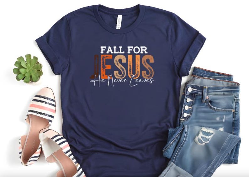 Puede incluir: Camiseta azul marino con el texto "FALL FOR JESUS He Never Leaves" en blanco, naranja y marr&oacute;n. La camiseta se combina con vaqueros azules y bailarinas a rayas. Tambi&eacute;n se ve una peque&ntilde;a planta suculenta verde.