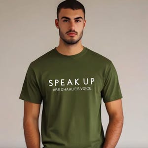 Puede incluir: Camiseta verde oliva con las palabras "SPEAK UP" y el hashtag "#BE CHARLIE'S VOICE" impresos en blanco. La camiseta tiene mangas cortas y cuello redondo.